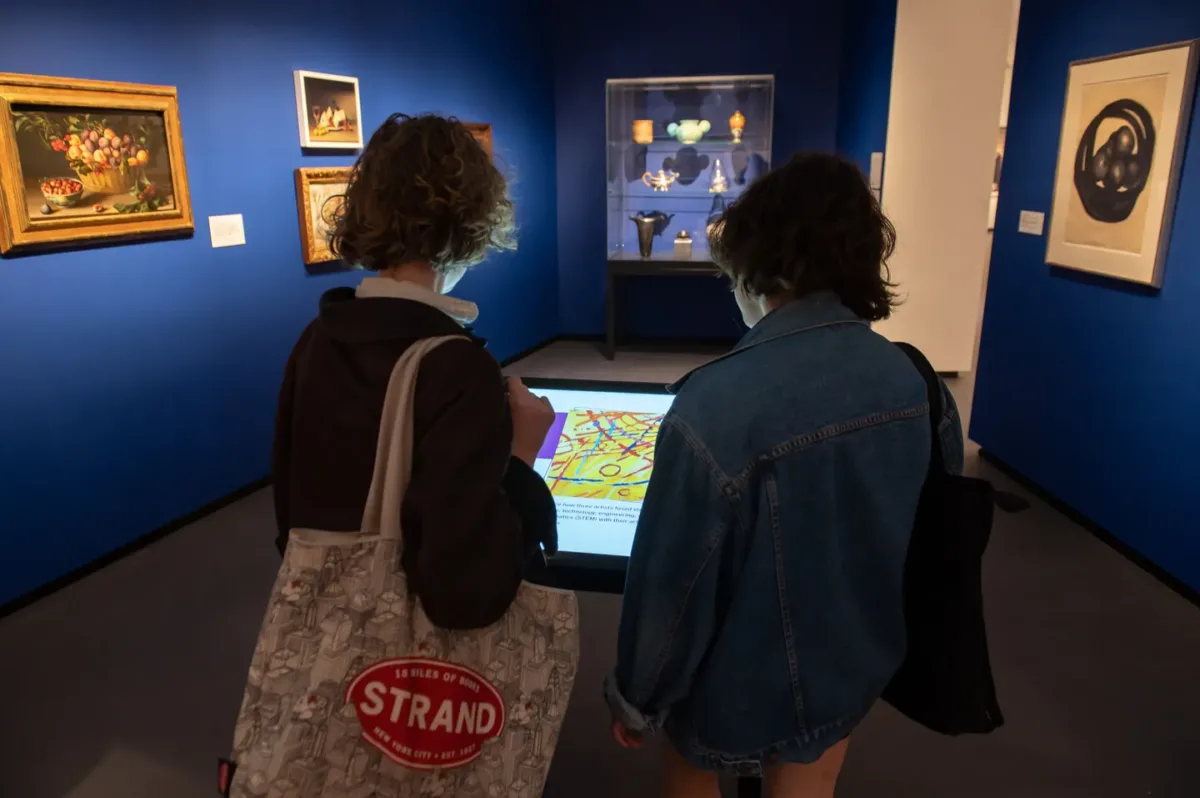 Two museum visitors are standing in a gallery. They are interacting with the digital screen in front of them, which shows different artworks.