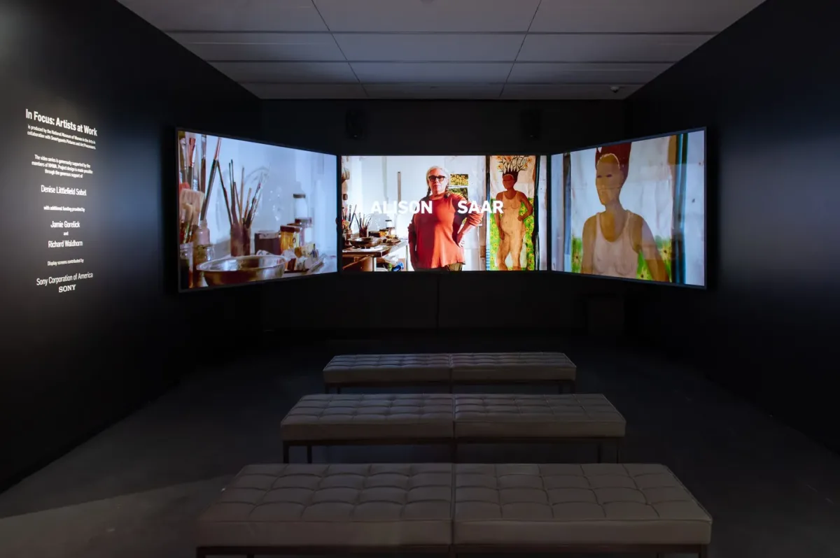 A dark gallery space with three large screens playing a video. Three tufted benches are in front of the screens. On the left wall, it says "In Focus: Artists at Work."