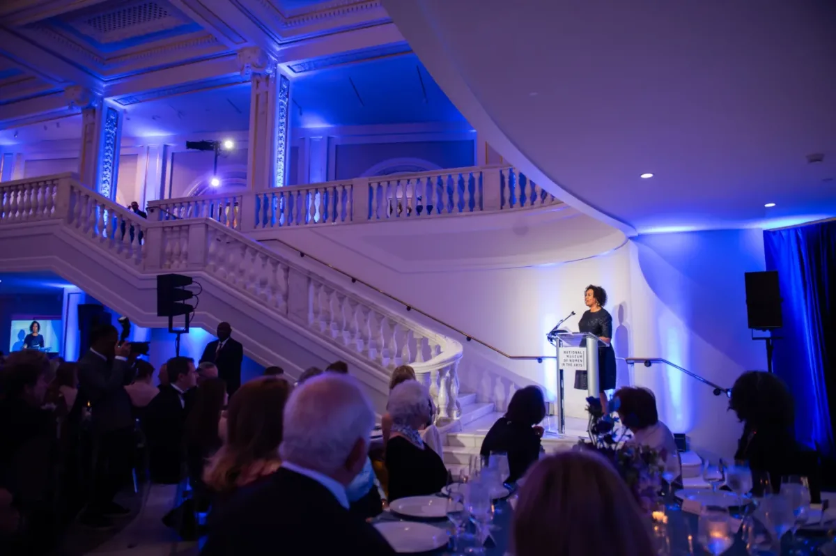 A medium-skinned woman with short brown hair wearing a long-sleeved, black gown stands behind a podium addressing a crowd of event attendees. The grand marble staircase behind her is lit for a formal evening event.