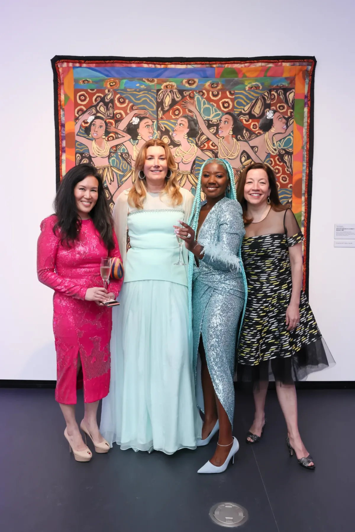 Four women with various skin tones, all in evening wear, pose for a picture together inside of an art gallery space.