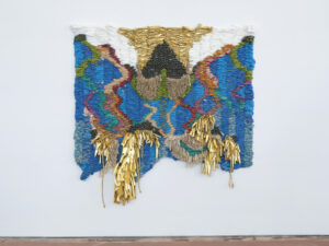 An abstract woven tapestry made with blue, green, red, pink, orange, white, and gold fabric. Small, round bells and gold cords adorn the tapestry.
