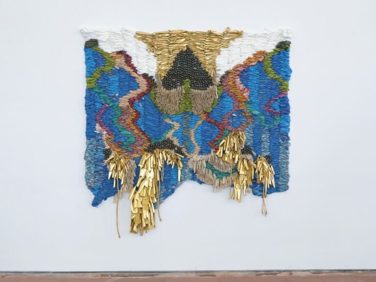 An abstract woven tapestry made with blue, green, red, pink, orange, white, and gold fabric. Small, round bells and gold cords adorn the tapestry.