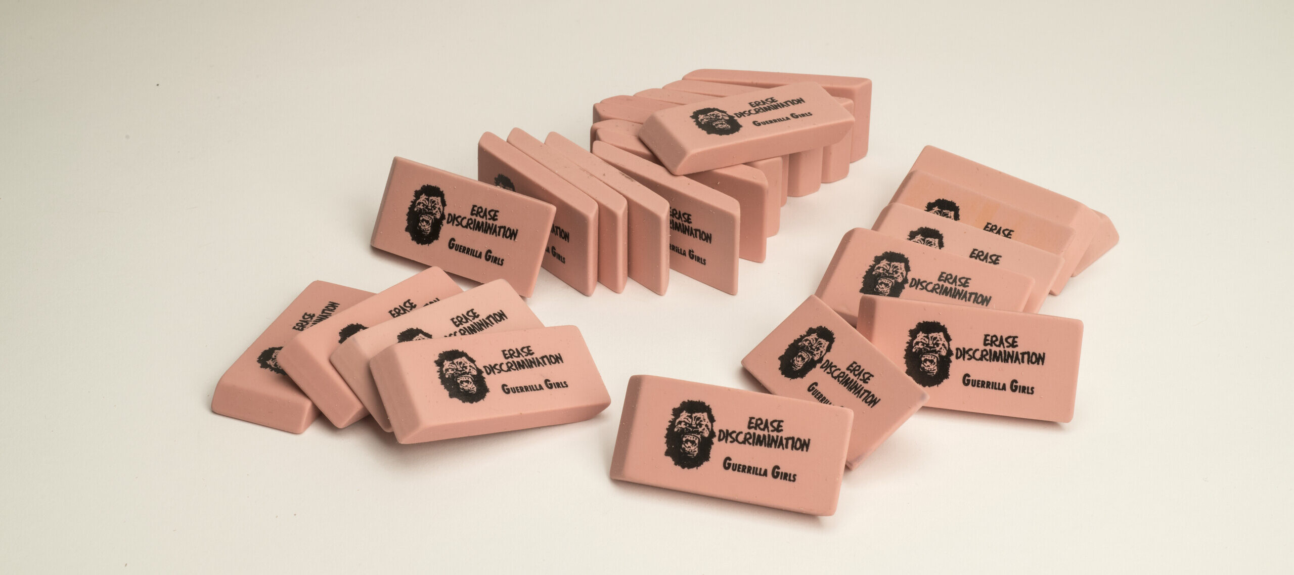 A pile of pink pencil erasers each with a screen print of a growling gorilla face mask, the command “ERASE DISCRIMINATION” in all capital letters, and the phrase “Guerrilla Girls,” which is the name of the troop of anonymous artists responsible for this work.