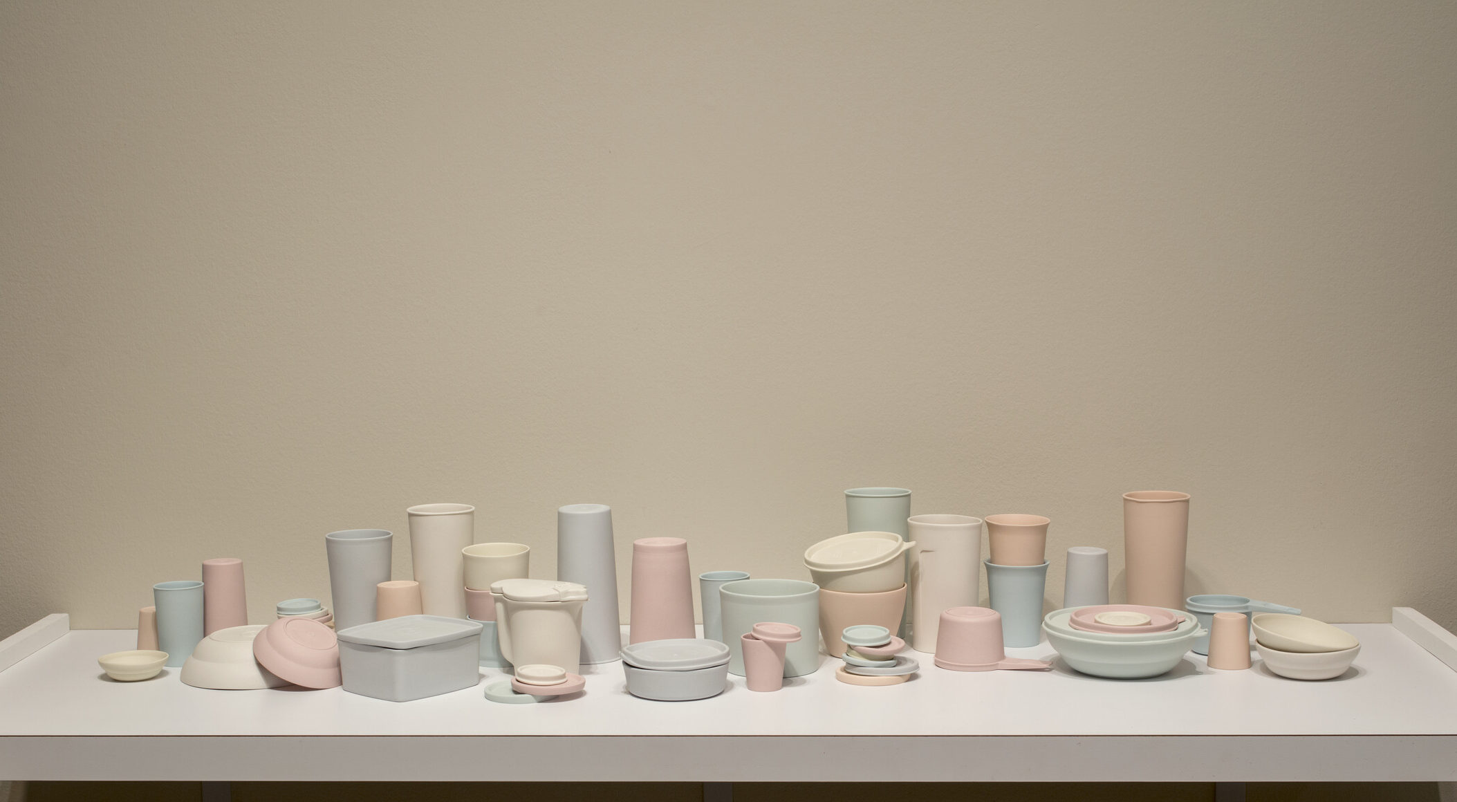 A collection of pastel-colored porcelain cups, bowls, containers, and lids that resemble plastic food containers. Some are stacked within and on top of each other. Some containers have lids firmly shut while on others the lids teeter off the edge of container rims.