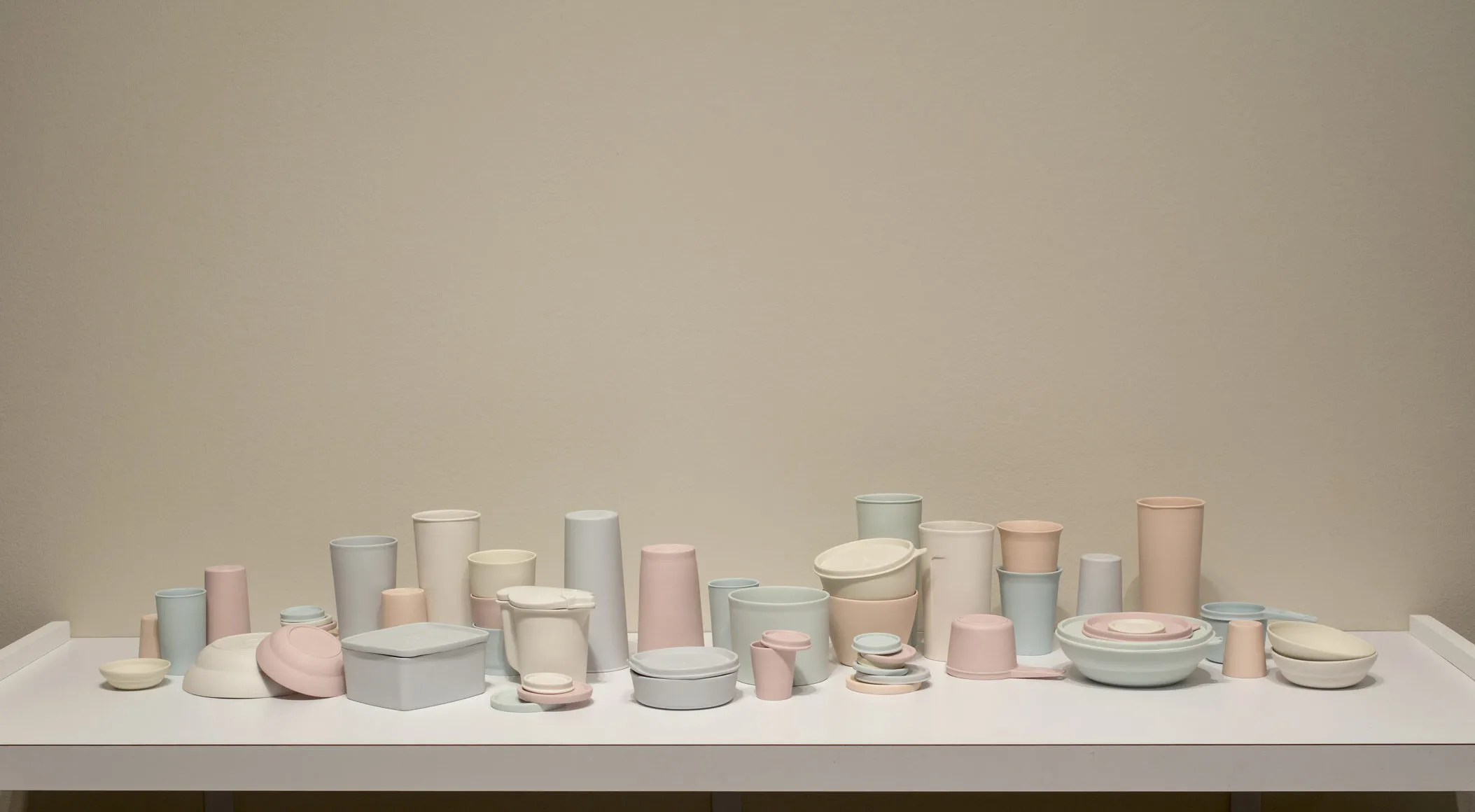 A collection of pastel-colored porcelain cups, bowls, containers, and lids that resemble plastic food containers. Some are stacked within and on top of each other. Some containers have lids firmly shut while on others the lids teeter off the edge of container rims.