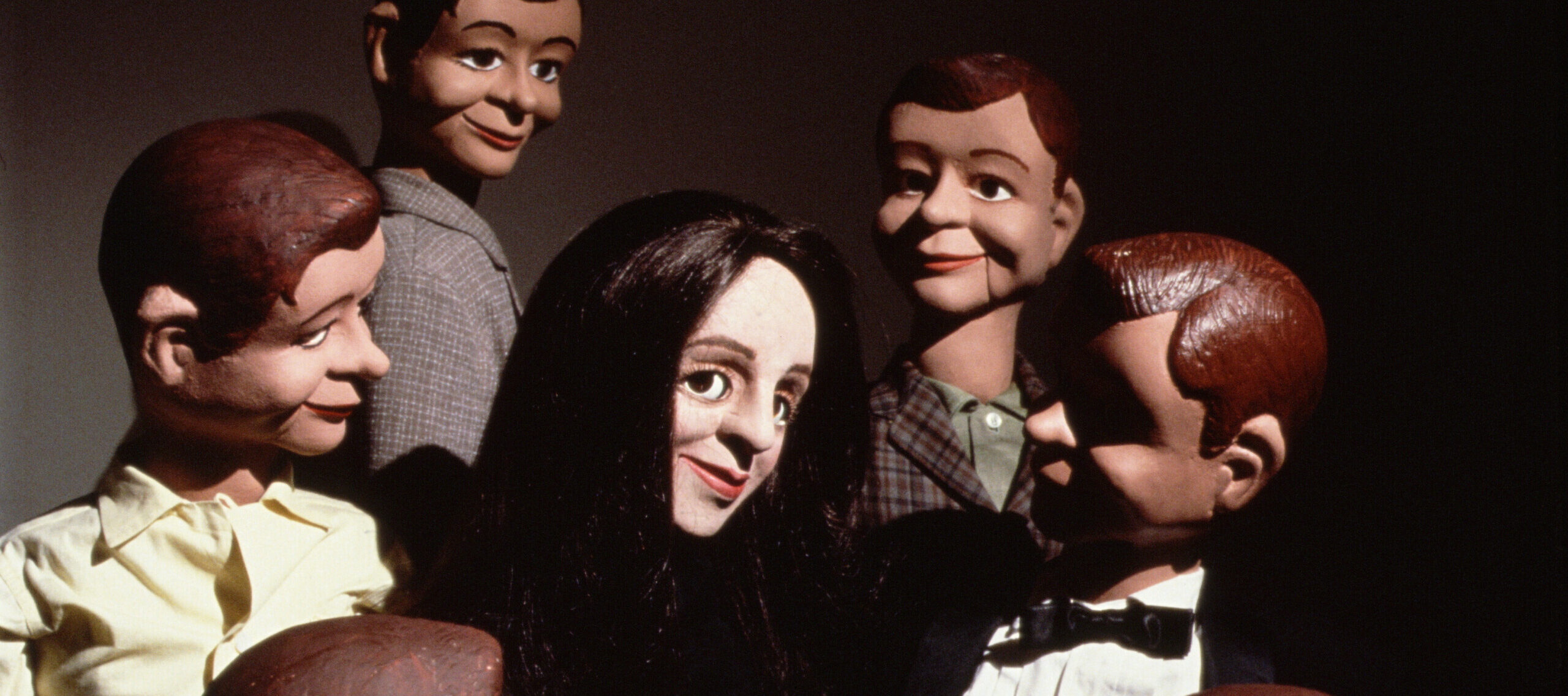 A light-skinned female ventriloquist dummy with straight black hair smiles in the center of a circle of six male dummy dolls with brown and dark hair.