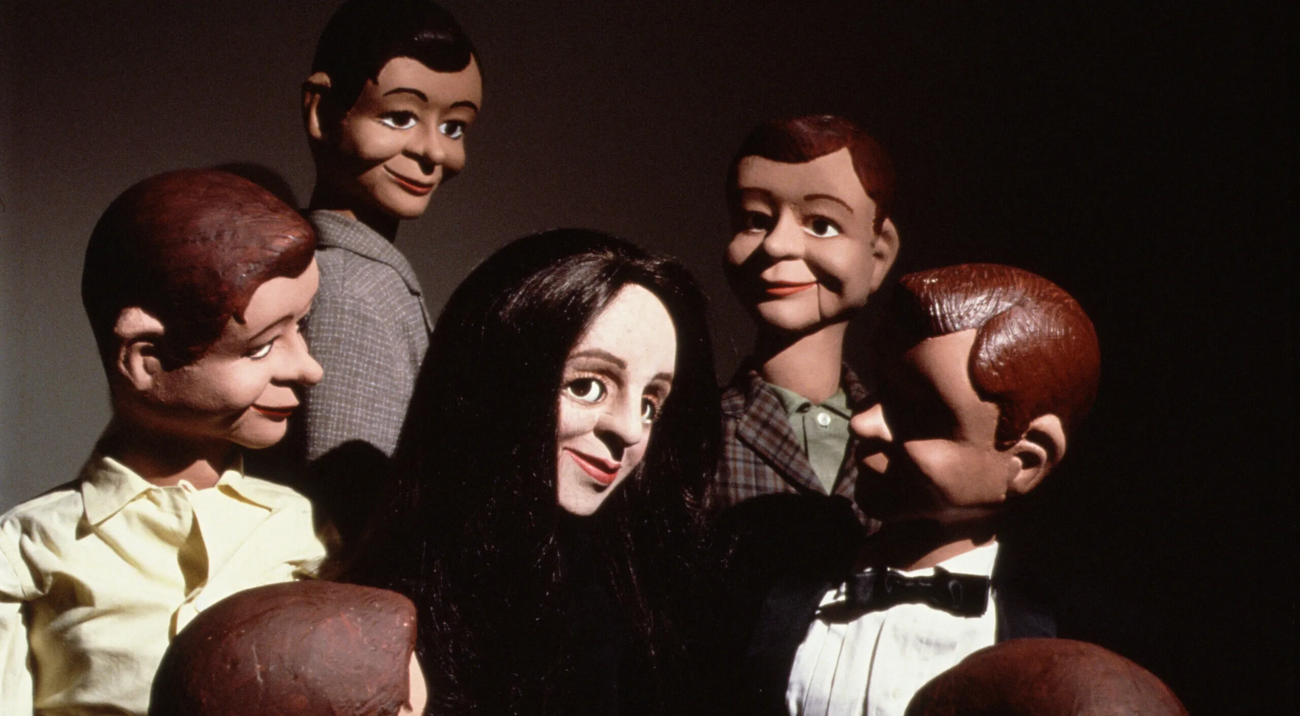 A light-skinned female ventriloquist dummy with straight black hair smiles in the center of a circle of six male dummy dolls with brown and dark hair.