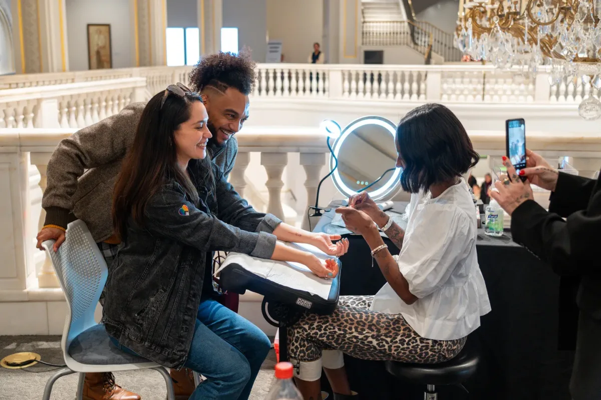 A woman in leopard printed pants and white top finishes giving a tattoo to a woman seated in front of her.