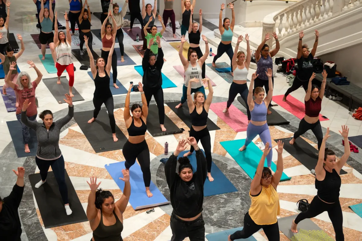 A crowd participates in a yoga class in the Great Hall of the National Museum of Women in the Arts. They all wear athletic apparel and are standing on their own yoga mats in a "warrior 2" pose.