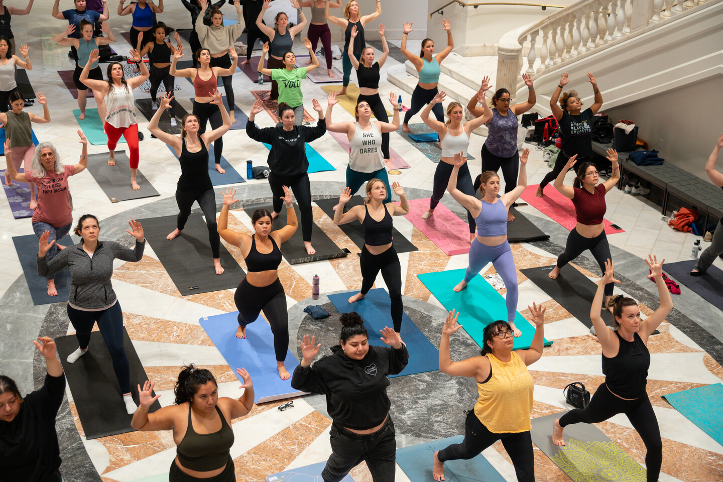 A crowd participates in a yoga class in the Great Hall of the National Museum of Women in the Arts. They all wear athletic apparel and are standing on their own yoga mats in a "warrior 2" pose.