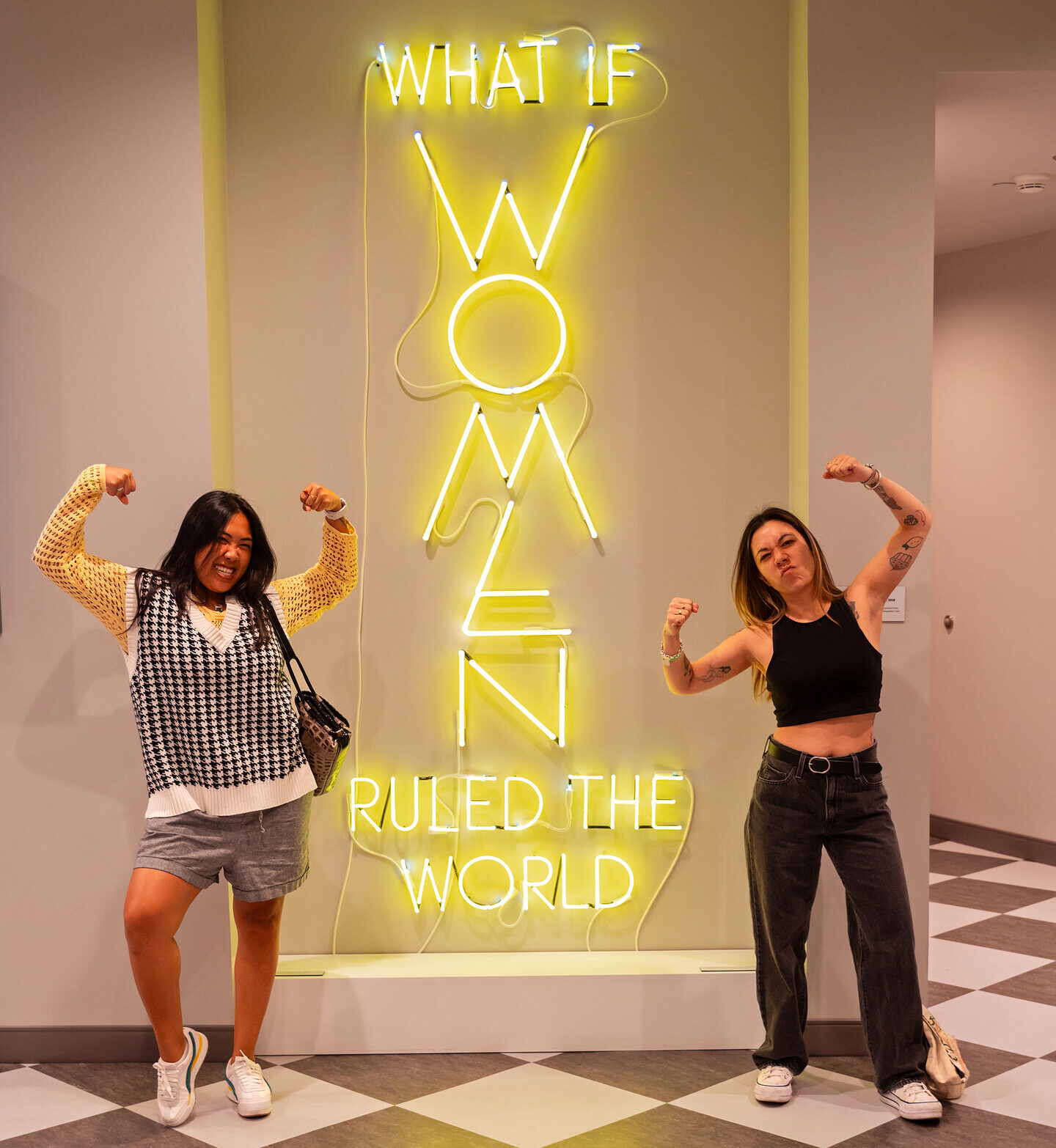 Two woman flex their arm muscles in front of a yellow neon sign that reads "what if women ruled the world"