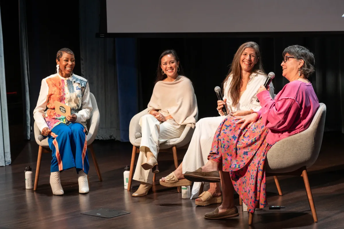 Four women sit on grey chairs on a black stage.