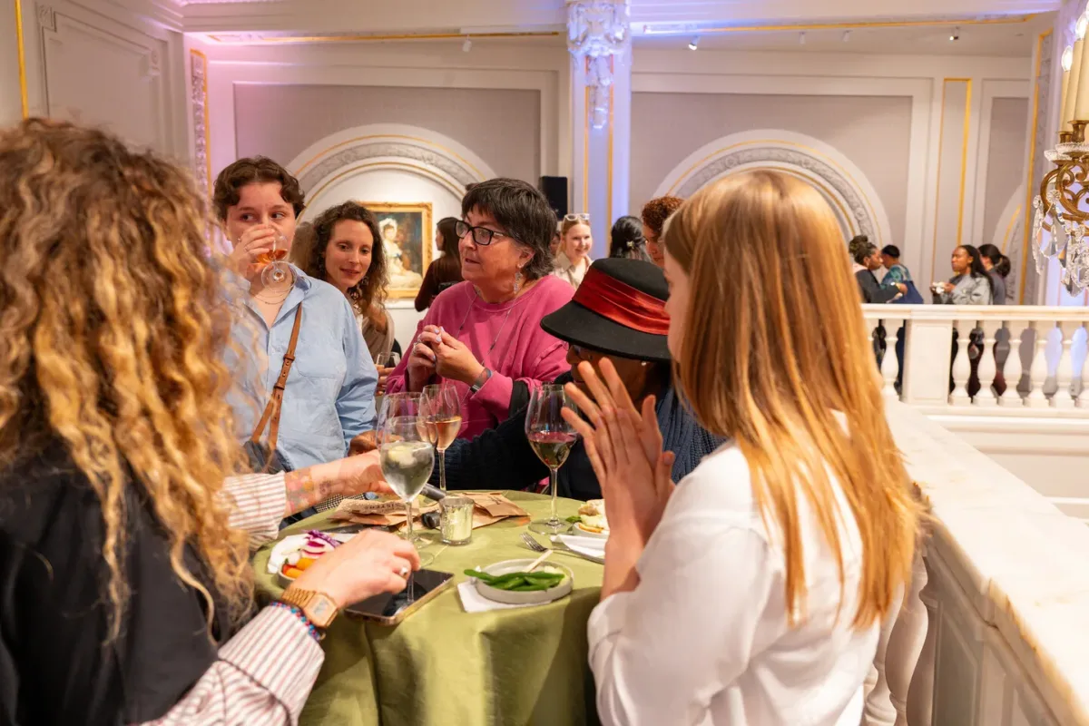 A woman with light colored skin and a pink top talks to four people around a green, circular table.