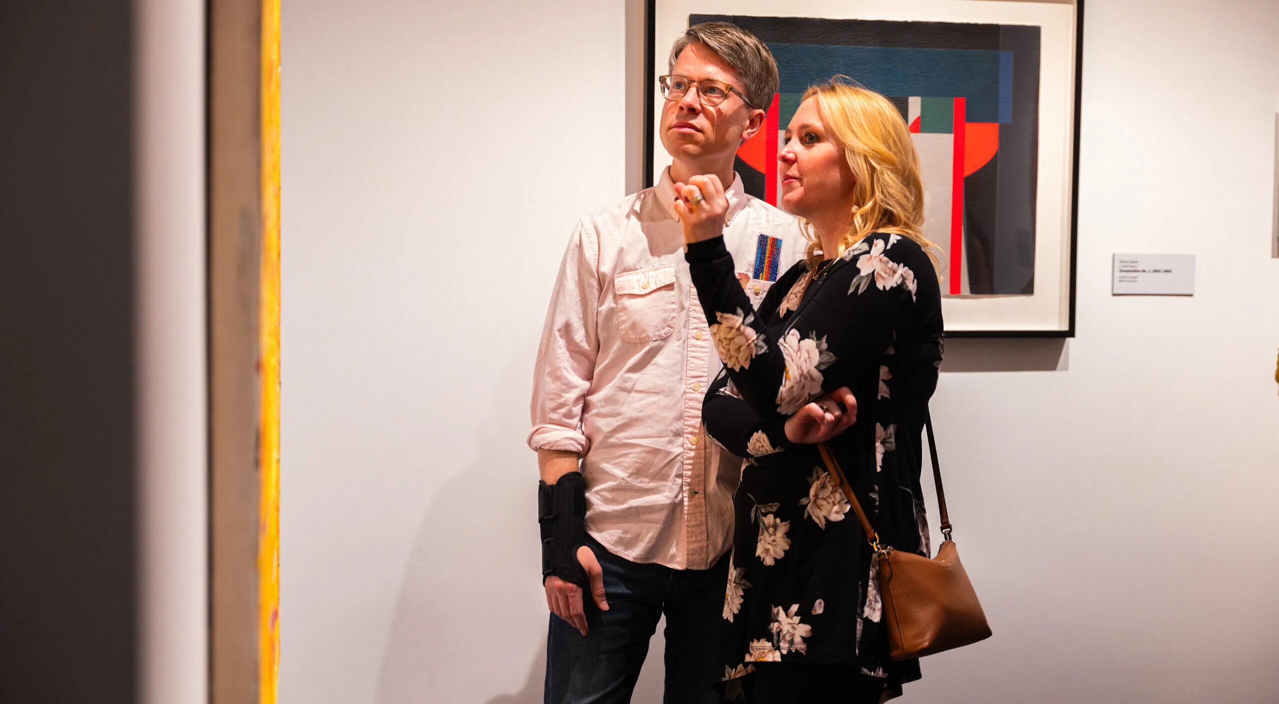 Two visitors with light skin look at a yellow painting. Behind them is a painting depicting a red, black, and gray image.