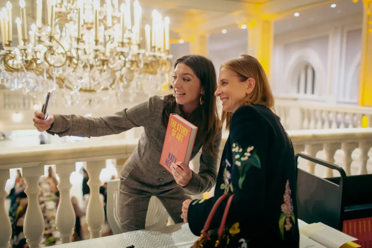 A woman with light colored skin, brown hair, and a grey jumpsuit holds an orange book whose cover reads "The Story of Art without Men, Katy Hessel." She leans over a table towards a woman with light colored skin and a floral sweater, and they pose to take a selfie on a phone.