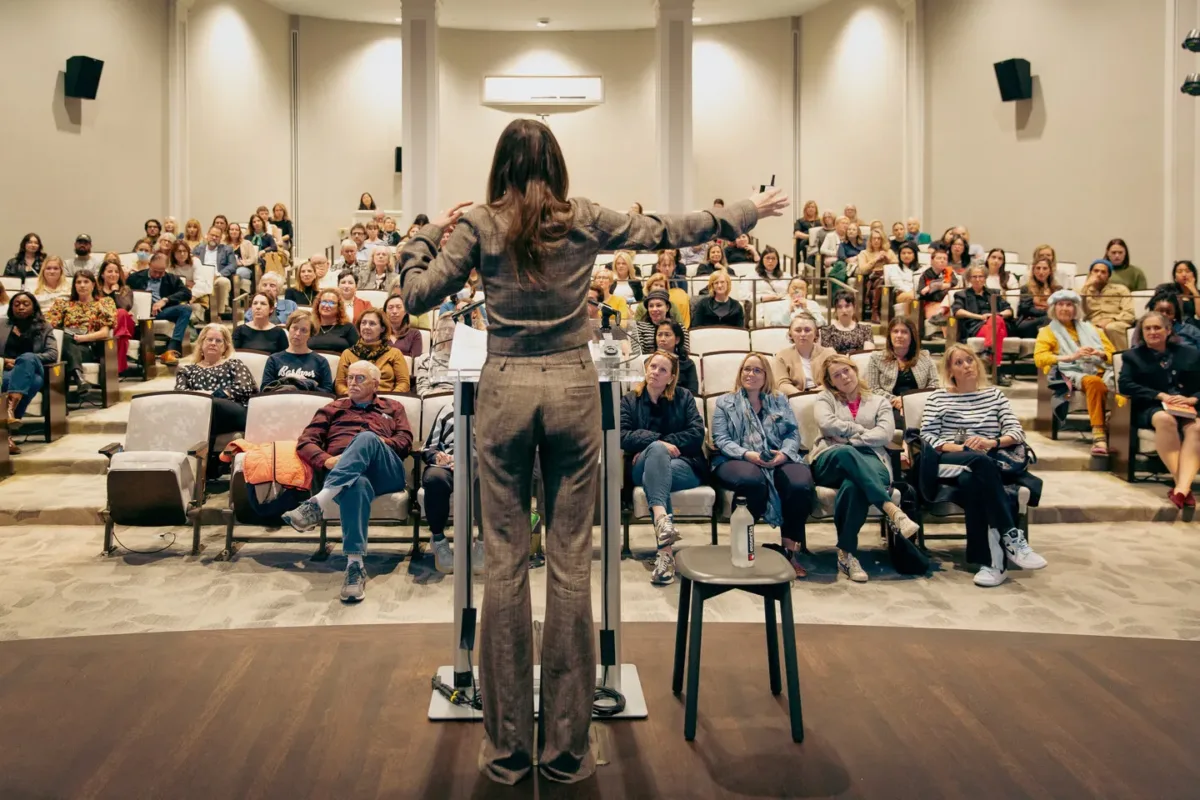 An audience of seated people watch woman with light colored skin and medium length brown hair and a grey jumpsuit speak behind a silver and clear podium.