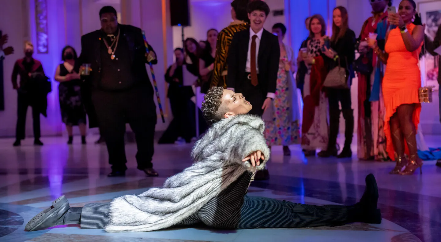 A drag king with a grey fur jacket and black pants does the splits in front of a crowd of people.