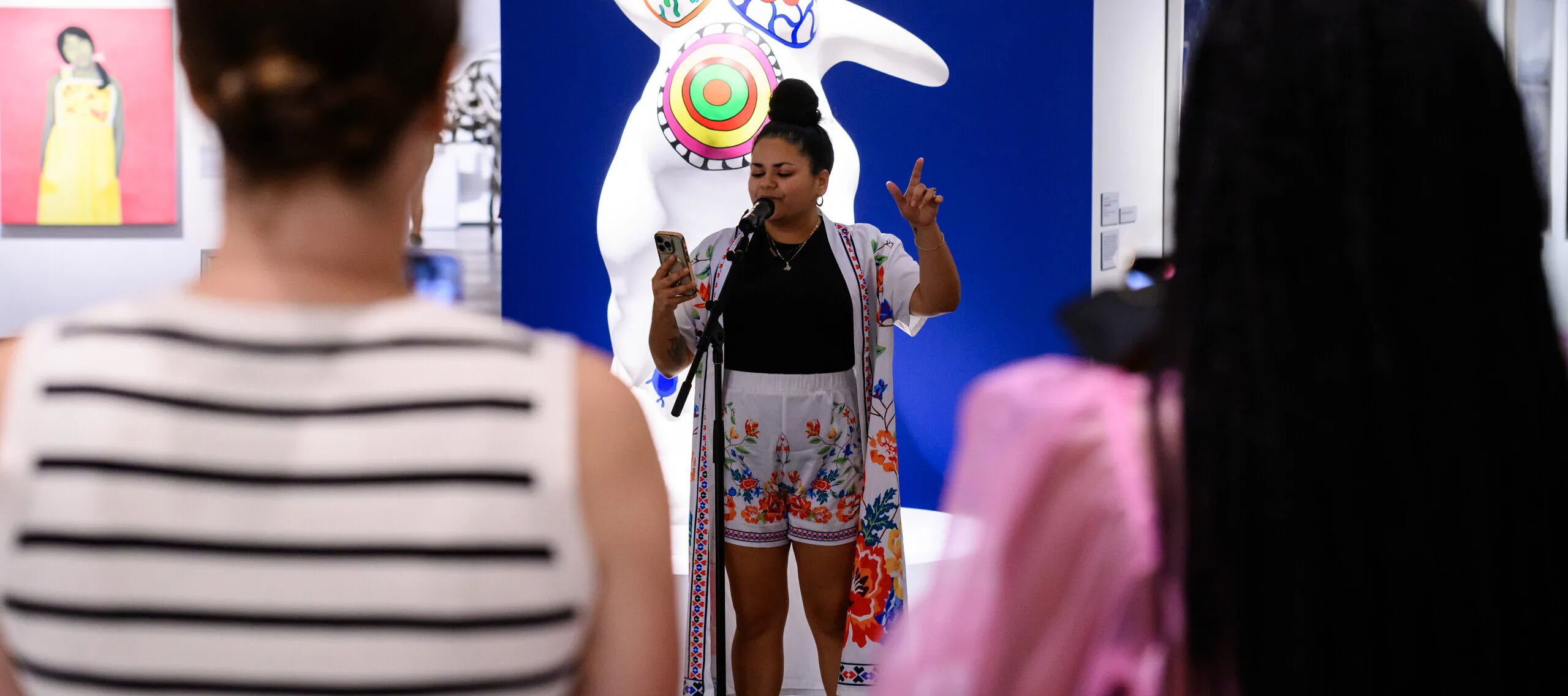 A woman performs into a microphone while standing in front of a statue of a woman's figure, surrounded by people in a gallery watching.