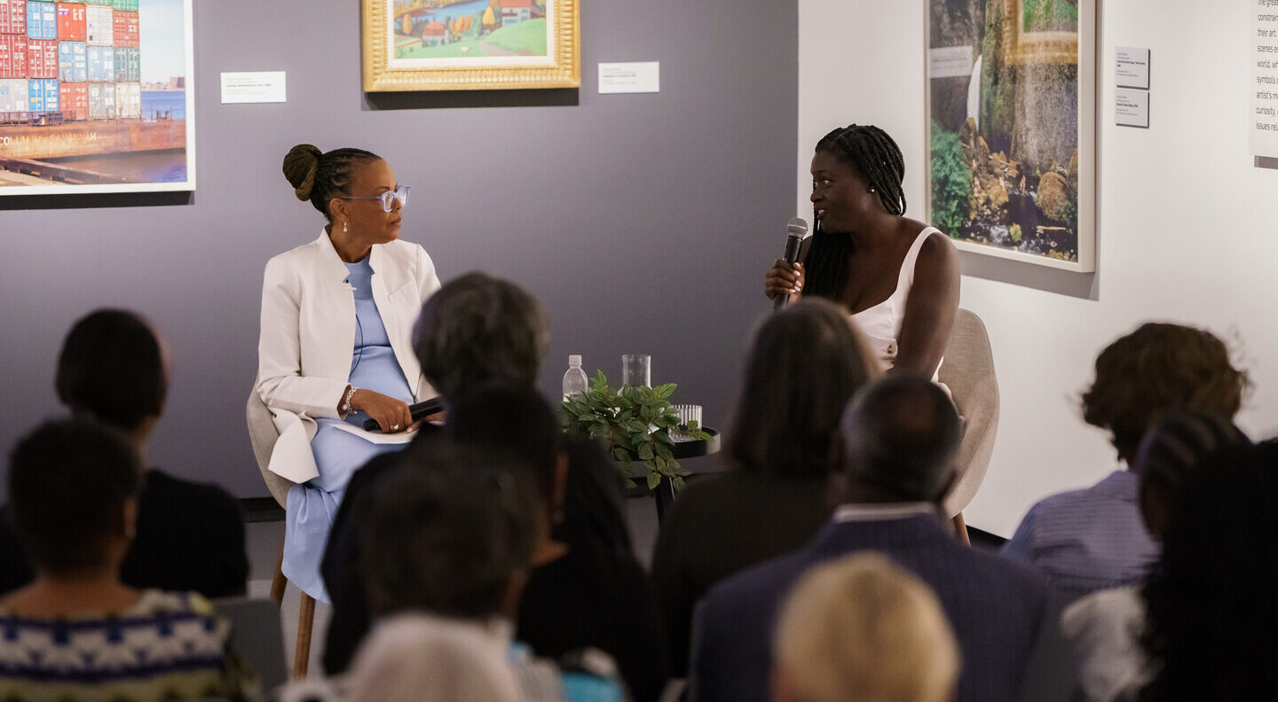 Two women with dark colored skin sit in chairs and talk to each other with microphones.