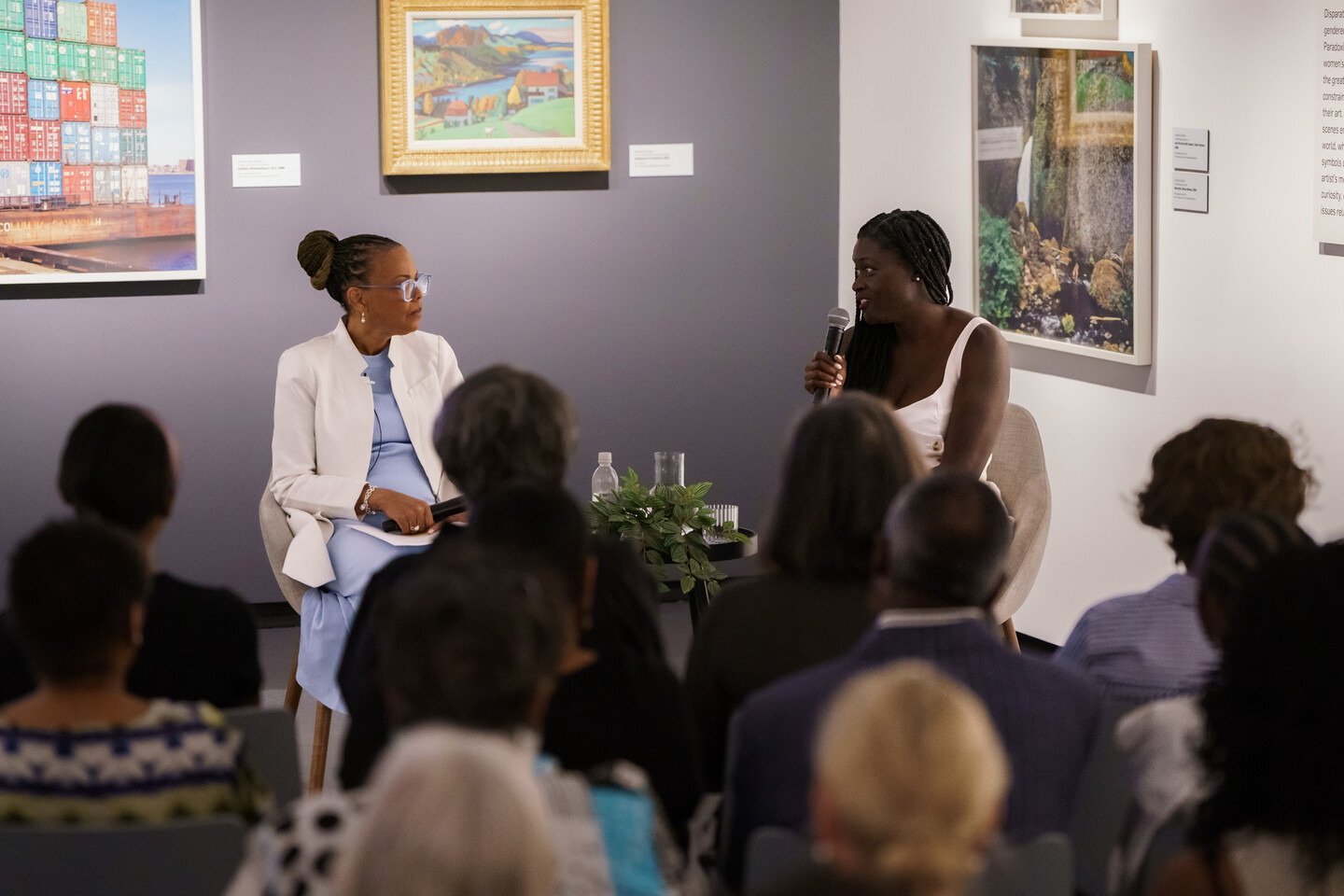 Two women with dark colored skin sit in chairs and talk to each other with microphones.