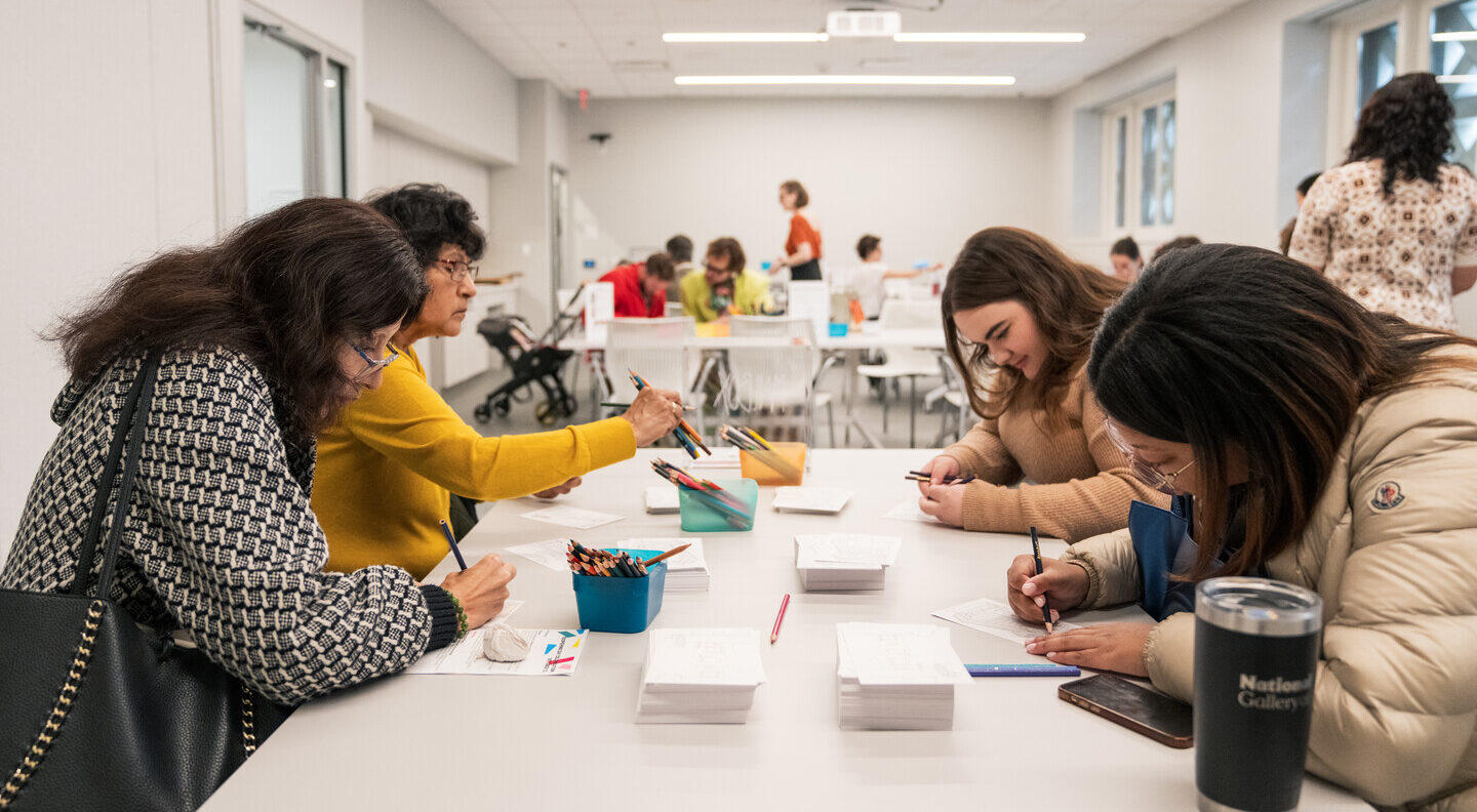 A group of women sit around a table and color on postcards with colored pencils.