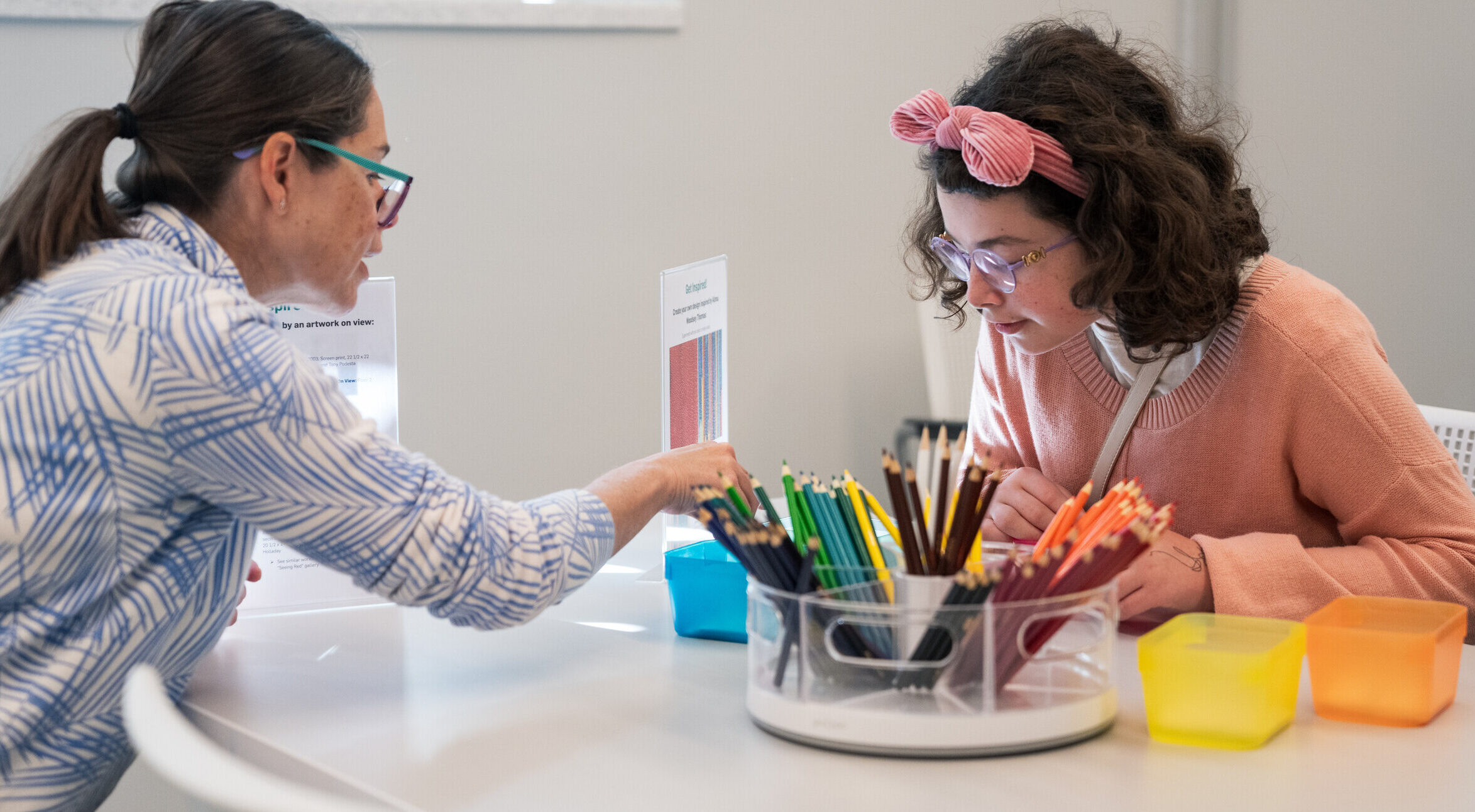 Two light-skinned women sit at a table and color with colored pencils.