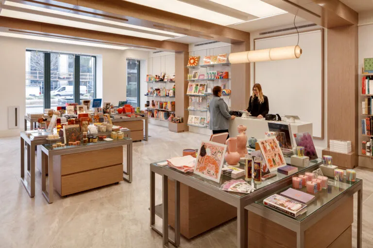 A museum gift shop is photographed at a wide angle. A colorful assortment of books, candles, textiles, and other small objects cover tables and shelves on the wall. A shopper is paying for items at a checkout table.