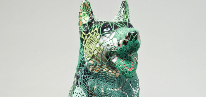 A mass-produced, ceramic German shepherd sits upright and alert with mouth open and tongue extended as if panting. Panels of elaborate hand-crafted crochet in shades of mint, citron, and emerald green form a skin-tight web that entirely envelops the dog.