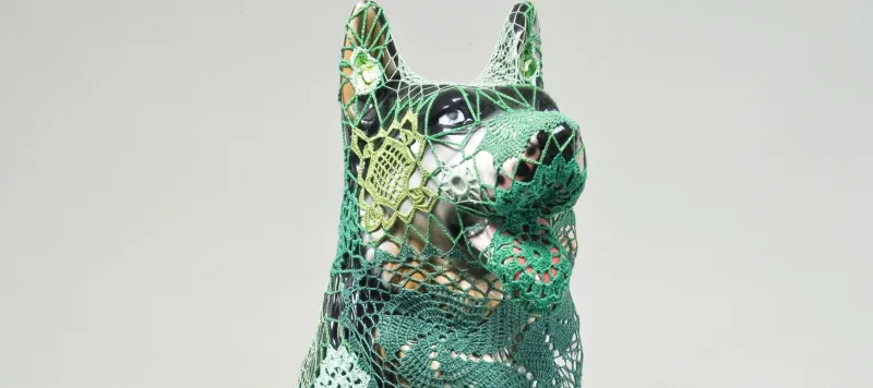 A mass-produced, ceramic German shepherd sits upright and alert with mouth open and tongue extended as if panting. Panels of elaborate hand-crafted crochet in shades of mint, citron, and emerald green form a skin-tight web that entirely envelops the dog.