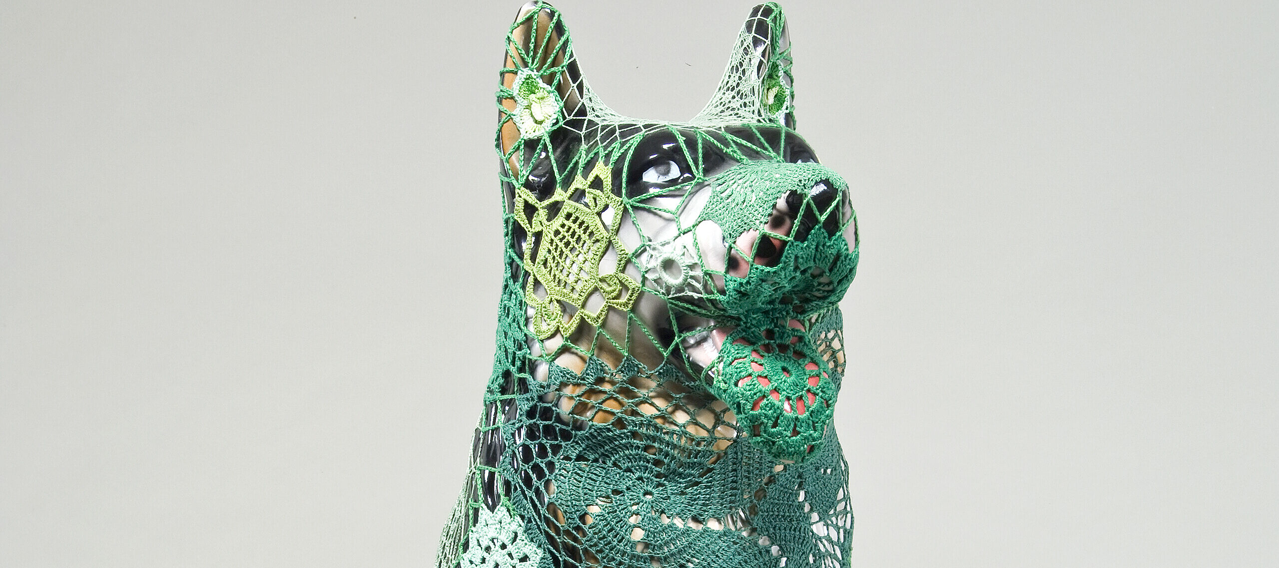 A mass-produced, ceramic German shepherd sits upright and alert with mouth open and tongue extended as if panting. Panels of elaborate hand-crafted crochet in shades of mint, citron, and emerald green form a skin-tight web that entirely envelops the dog.