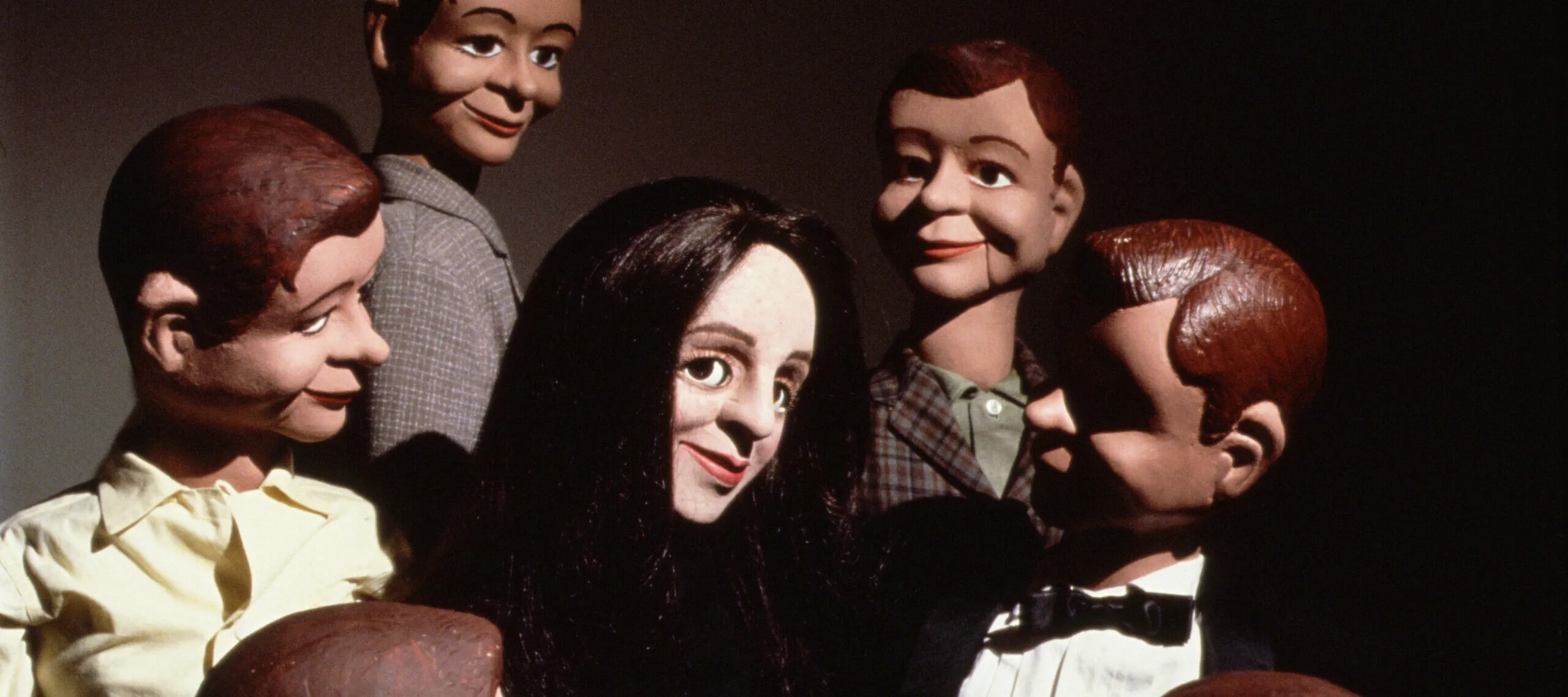 A light-skinned female ventriloquist dummy with straight black hair smiles in the center of a circle of six male dummy dolls with brown and dark hair.