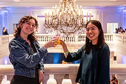 Two women stand in front of a white marble railing and tap their drinks together, smiling at the camera.