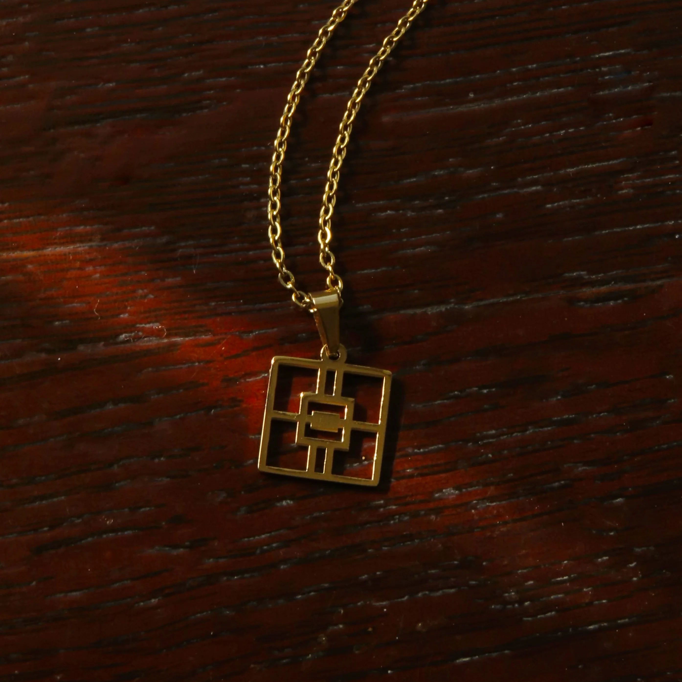 A gold necklace sits atop a wood base with one singular ray of sunshine crossing it. It is a simple square, split into four squares with a central square and fill, reminiscent of a decorative window. It has a long gold chain.