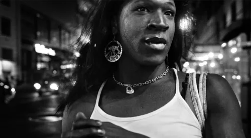 A black-and-white close-up image shows the face and upper body of a dark-skinned woman as she gazes off to the side. She wears a sleeveless white shirt, chain necklace with heart pendant, and large round earrings inscribed with the word “TAURUS.”