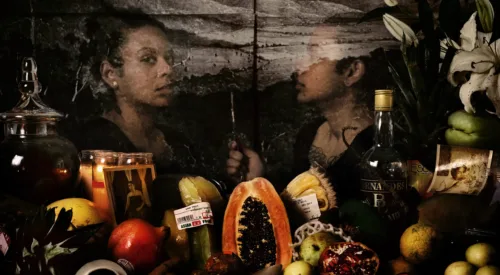 An assortment of flowers, colorful fruits, glass bottles, and photographs are visible in front of a transparent image of a dark-skinned woman who looks out at the viewer while her reflection looks at her. Behind her is a black-and-white image of a mountainous landscape.