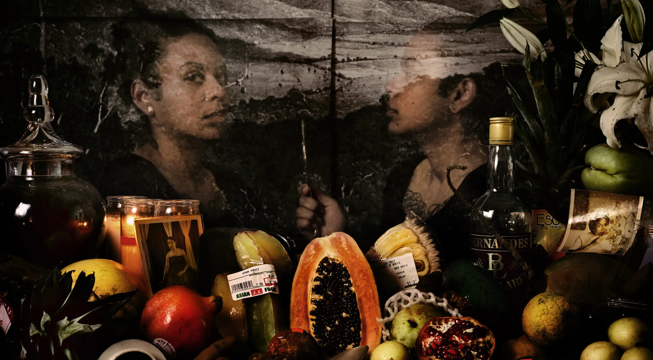 An assortment of flowers, colorful fruits, glass bottles, and photographs are visible in front of a transparent image of a dark-skinned woman who looks out at the viewer while her reflection looks at her. Behind her is a black-and-white image of a mountainous landscape.