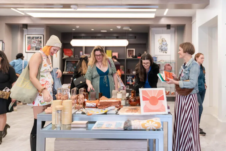 A group of women are standing in a shop around a table with colorful books, prints, and other objects on it. They have light skin tones and are wearing colorful dresses.