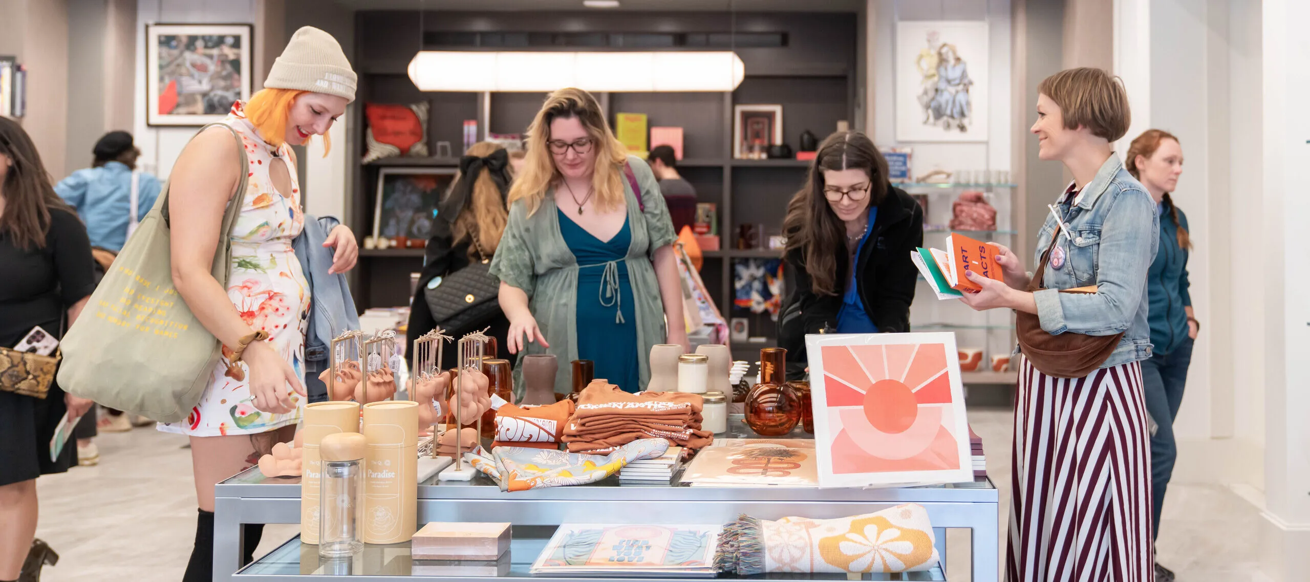 A group of women are standing in a shop around a table with colorful books, prints, and other objects on it. They have light skin tones and are wearing colorful dresses.