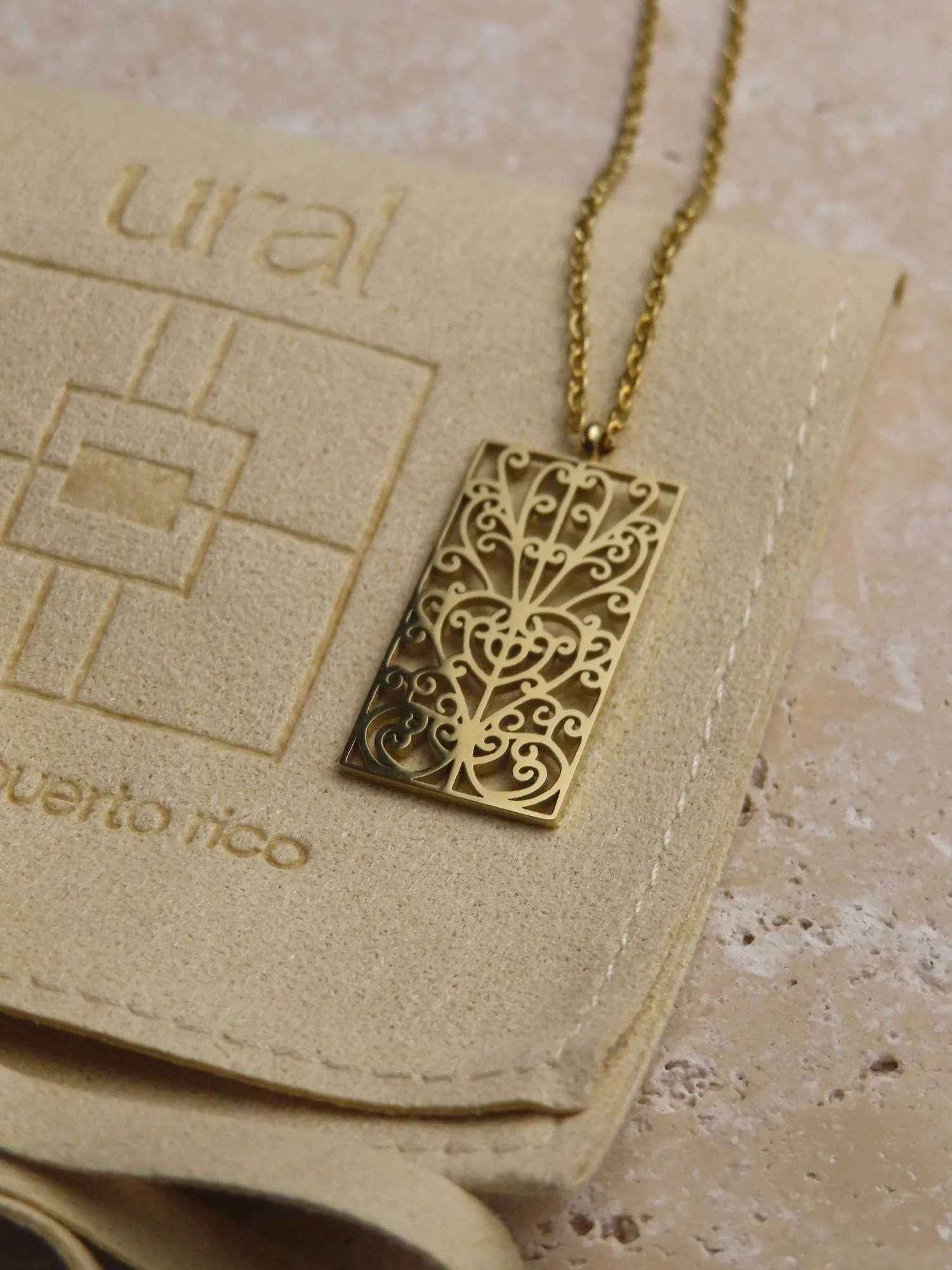 A gold rectangular pendant necklace features a central design of curves and loops that form a heart. It is reminiscent of iron work on the outer doors of some houses in Latin America and the Caribbean.