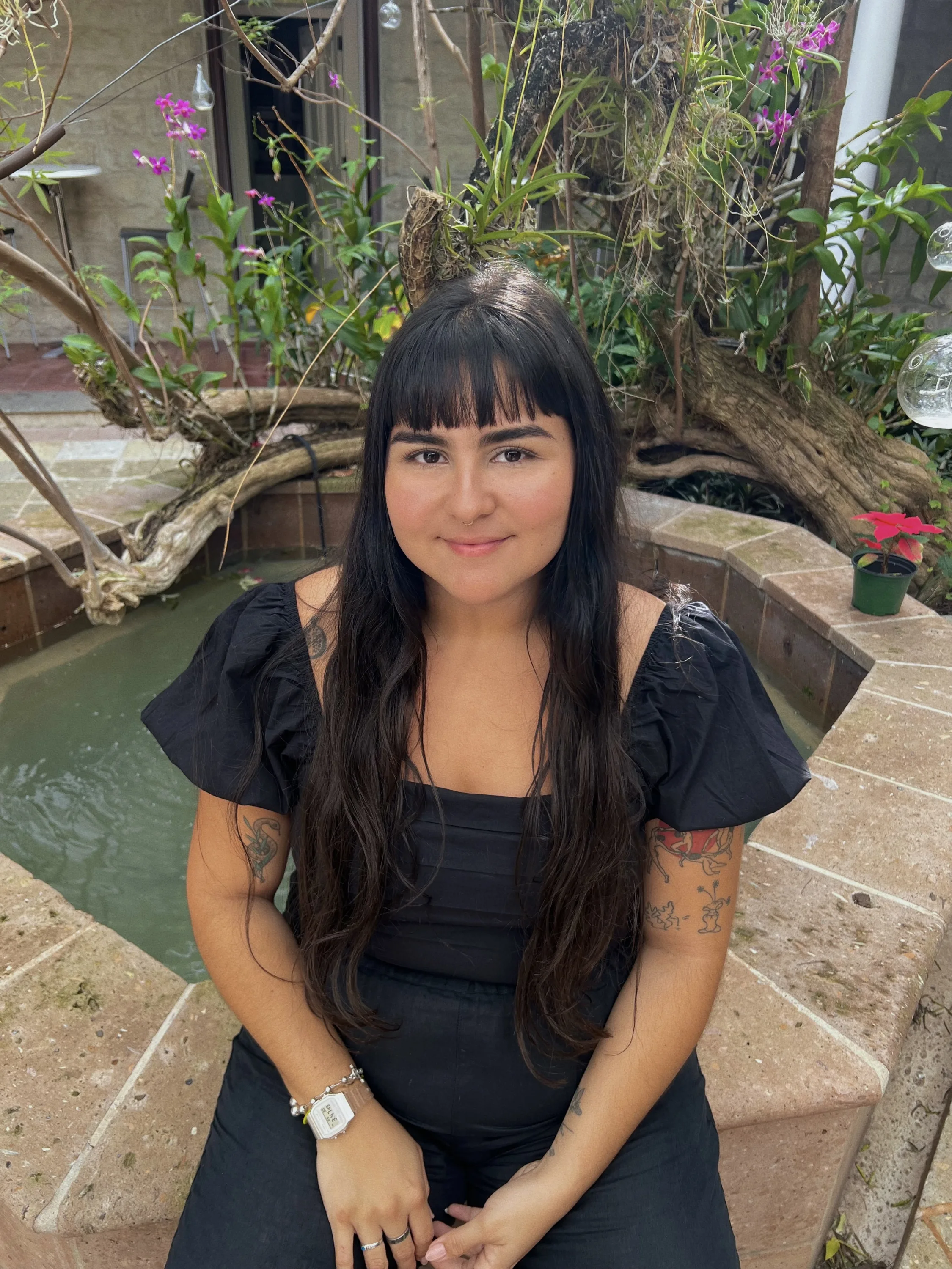 A young woman with medium-light skin tone and long, straight black hair and bangs sits on the stone ledge of a courtyard fountain. She wears a black dress with ruffle sleeves. Some tattoos are visible on her arms. She smiles warmly at the camera. Behind her is a tree, growing to the left and sprouting some green leaves and small purple flowers.