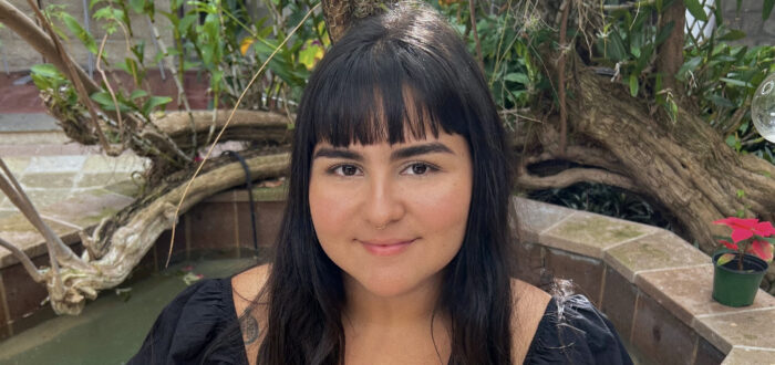 A young woman with medium-light skin tone and long, straight black hair and bangs sits on the stone ledge of a courtyard fountain. She wears a black dress with ruffle sleeves. Some tattoos are visible on her arms. She smiles warmly at the camera. Behind her is a tree, growing to the left and sprouting some green leaves and small purple flowers.