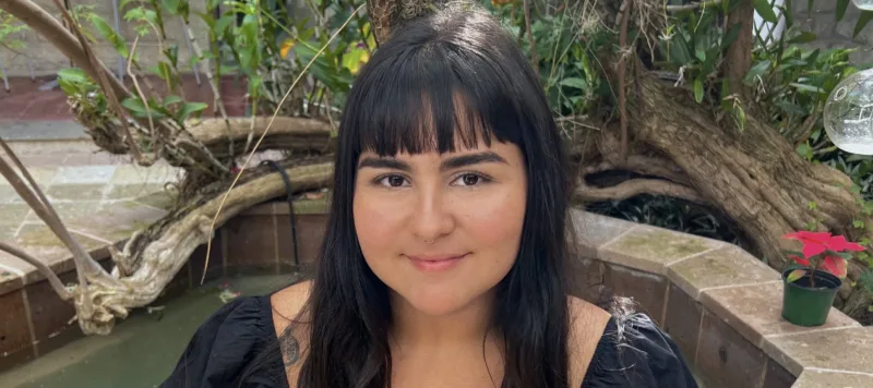 A young woman with medium-light skin tone and long, straight black hair and bangs sits on the stone ledge of a courtyard fountain. She wears a black dress with ruffle sleeves. Some tattoos are visible on her arms. She smiles warmly at the camera. Behind her is a tree, growing to the left and sprouting some green leaves and small purple flowers.