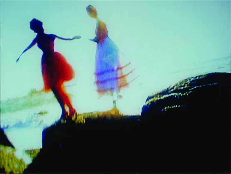 A blurry image of two figures wearing dresses, appearing to be standing on a large rock or outcropping. Their arms are aloft as in mid-dance or stride.