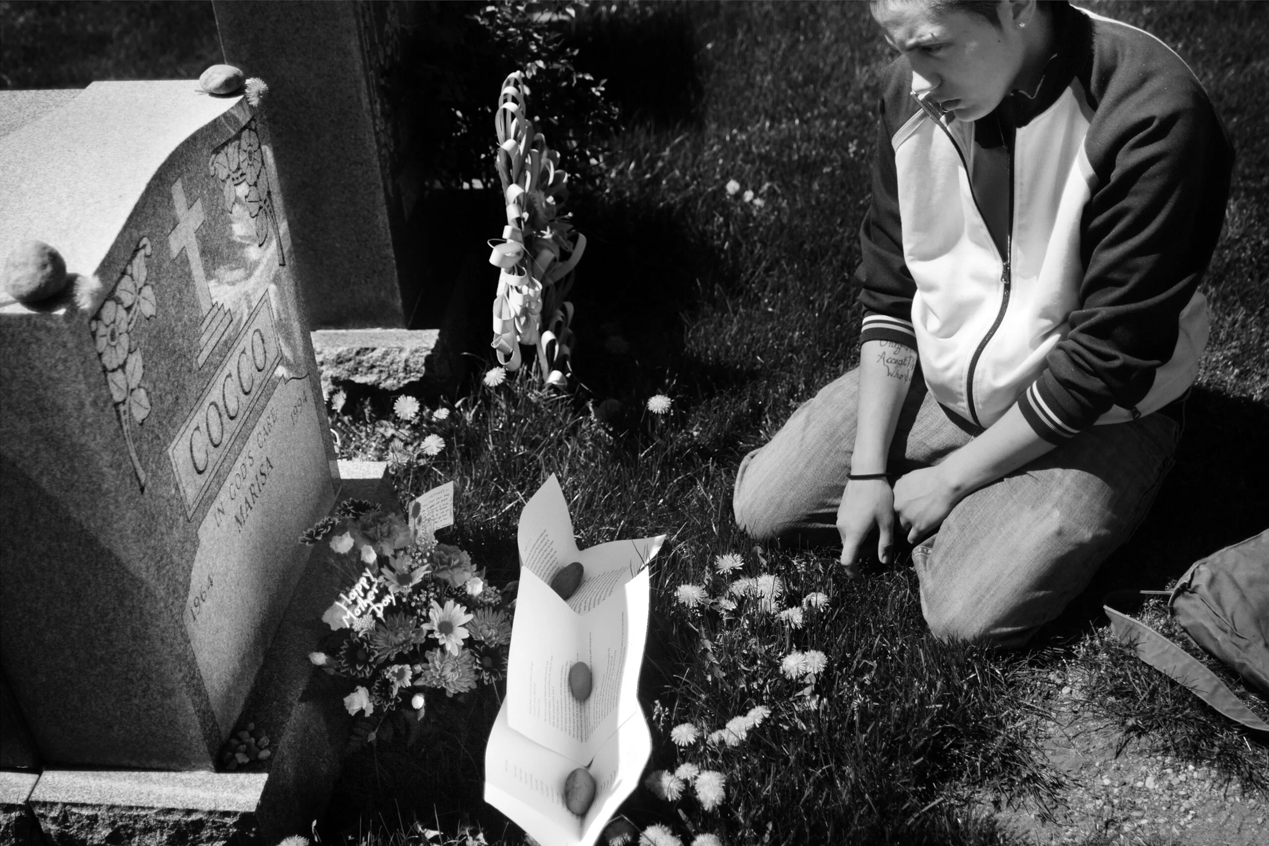 A black-and-white image shows a light-complexioned person kneeling by a gravestone marked with a cross and the word “COCCO.” In front of the gravestone, flowers and three sheets of paper held down with stones lie on the ground. NEEDS CURATORIAL APPRO