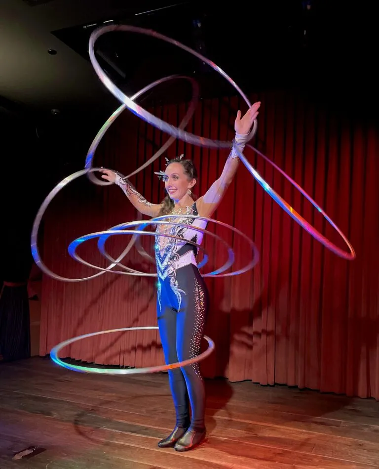 A performer stands onstage with six hula hoops in motion around their body.