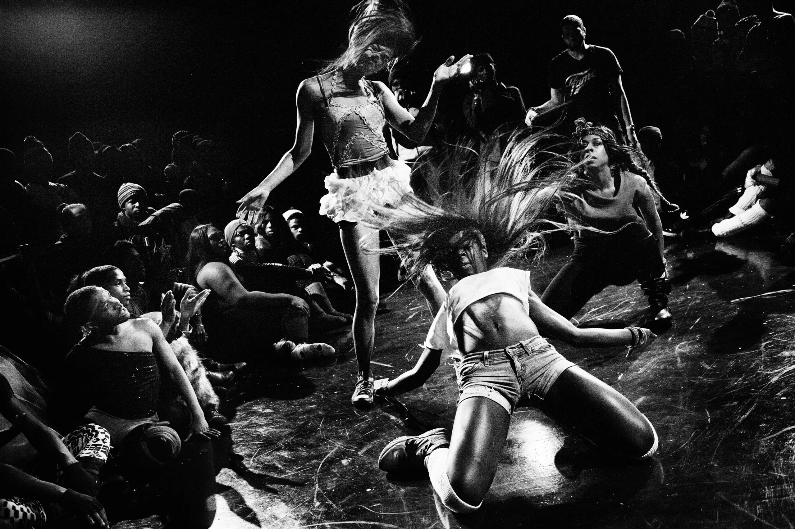 A black-and-white image shows a group of figures during a performance. The person in the foreground wears shorts and cropped shirt; they kneel on the ground with flailing hair while another person in a white skirt bends her head to one side. Other people