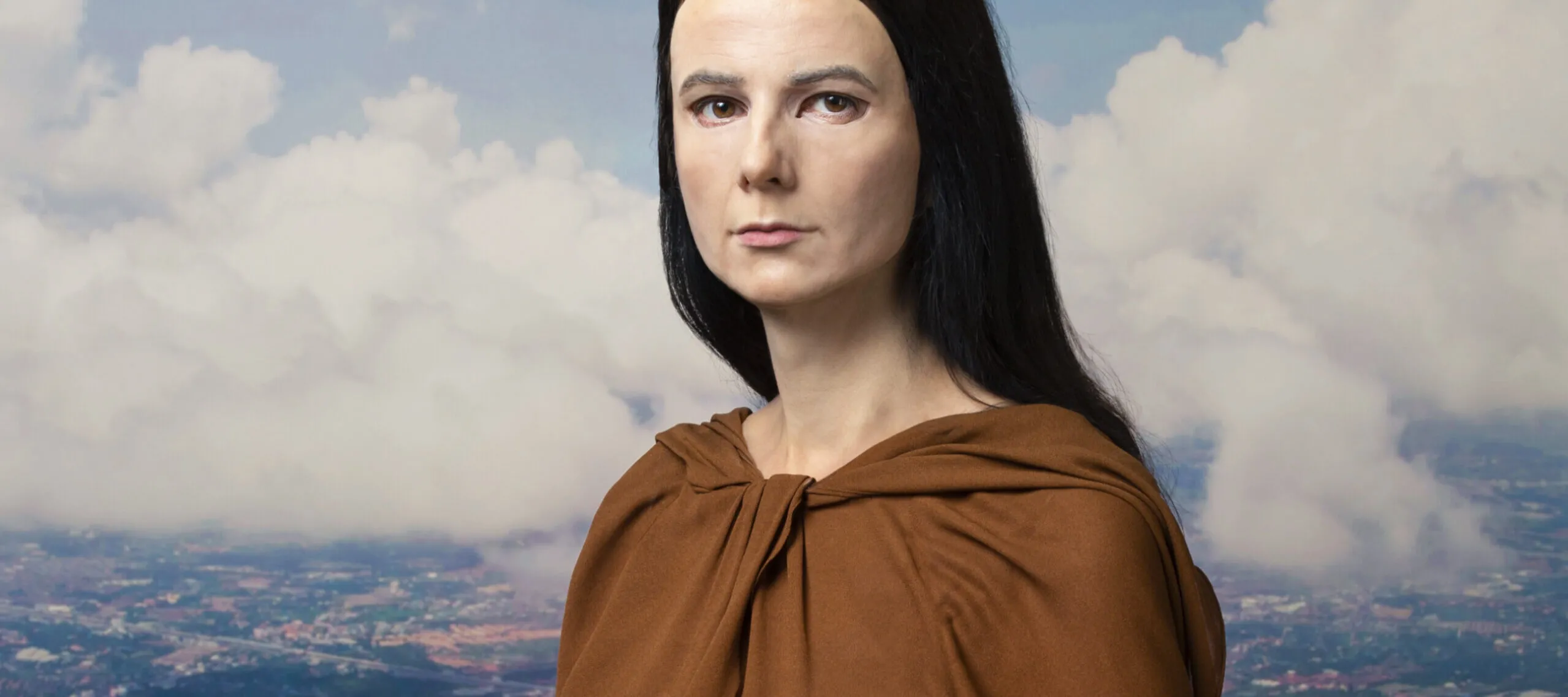 A light-skinned woman wears a realistic mask of another woman with the same skin tone. She sits posed like the Mona Lisa. She has long dark hair and wears a brown dress. The backdrop behind her shows  a cityscape with rivers and clouds.