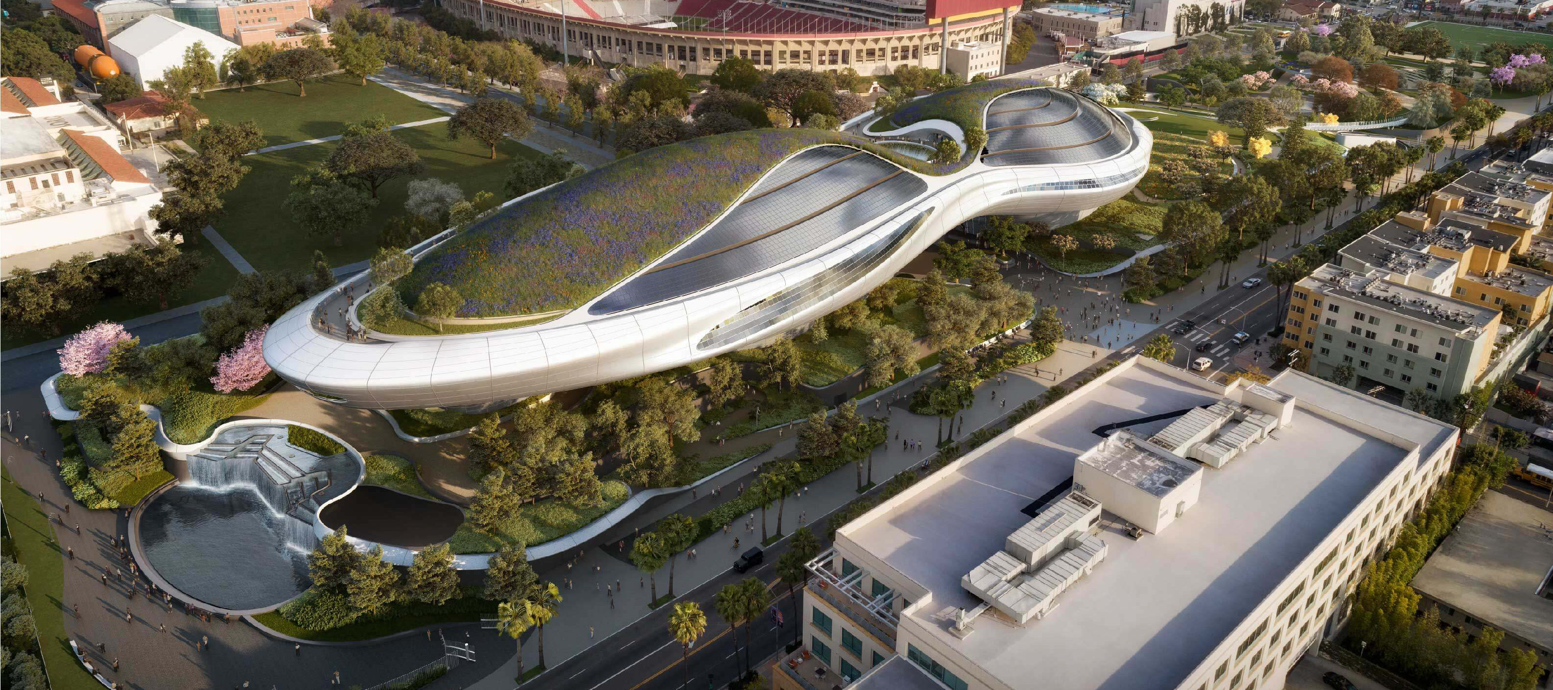 An aerial view of an elliptical shaped futuristic building that has grass and trees on top; the building is surrounded by a green park in a city with a football stadium behind it.