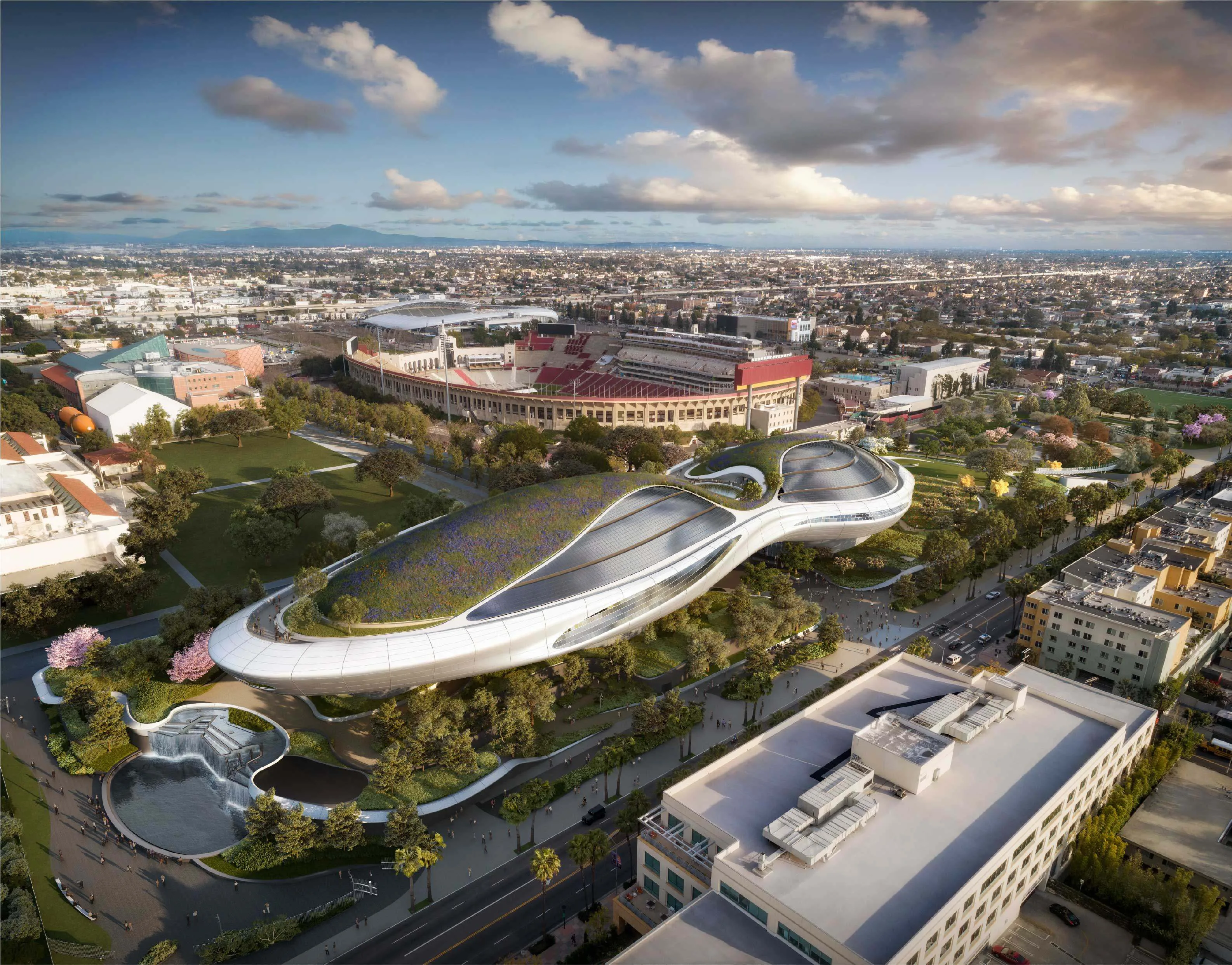 An aerial view of an elliptical shaped futuristic building that has grass and trees on top; the building is surrounded by a green park in a city with a football stadium behind it.