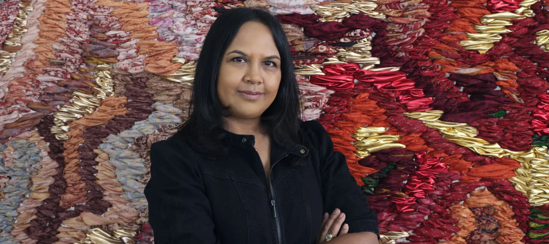 A photograph of a medium-skinned woman with black hair, shown from the waist up. She is wearing a black zip-up jacket and has her arms crossed in front of her. She stands in front of a large-scale abstract tapestry made of red, orange, pink, and gold fabric.